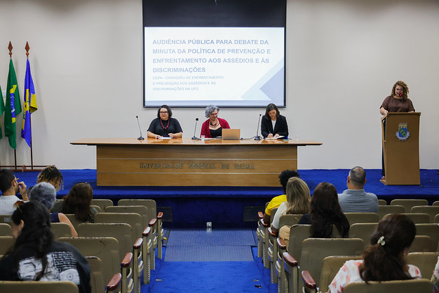 Na quarta-feira (14), o auditório da Reitoria recebeu a audiência pública para discussão da minuta da Política de Enfrentamento e Prevenção aos Assédios e Discriminações no âmbito da UFC (Foto: Ribamar Net/UFC) Imagem: três mulheres sentadas em uma mesa e outra mulher falando no púlpito do auditório da Reitoria