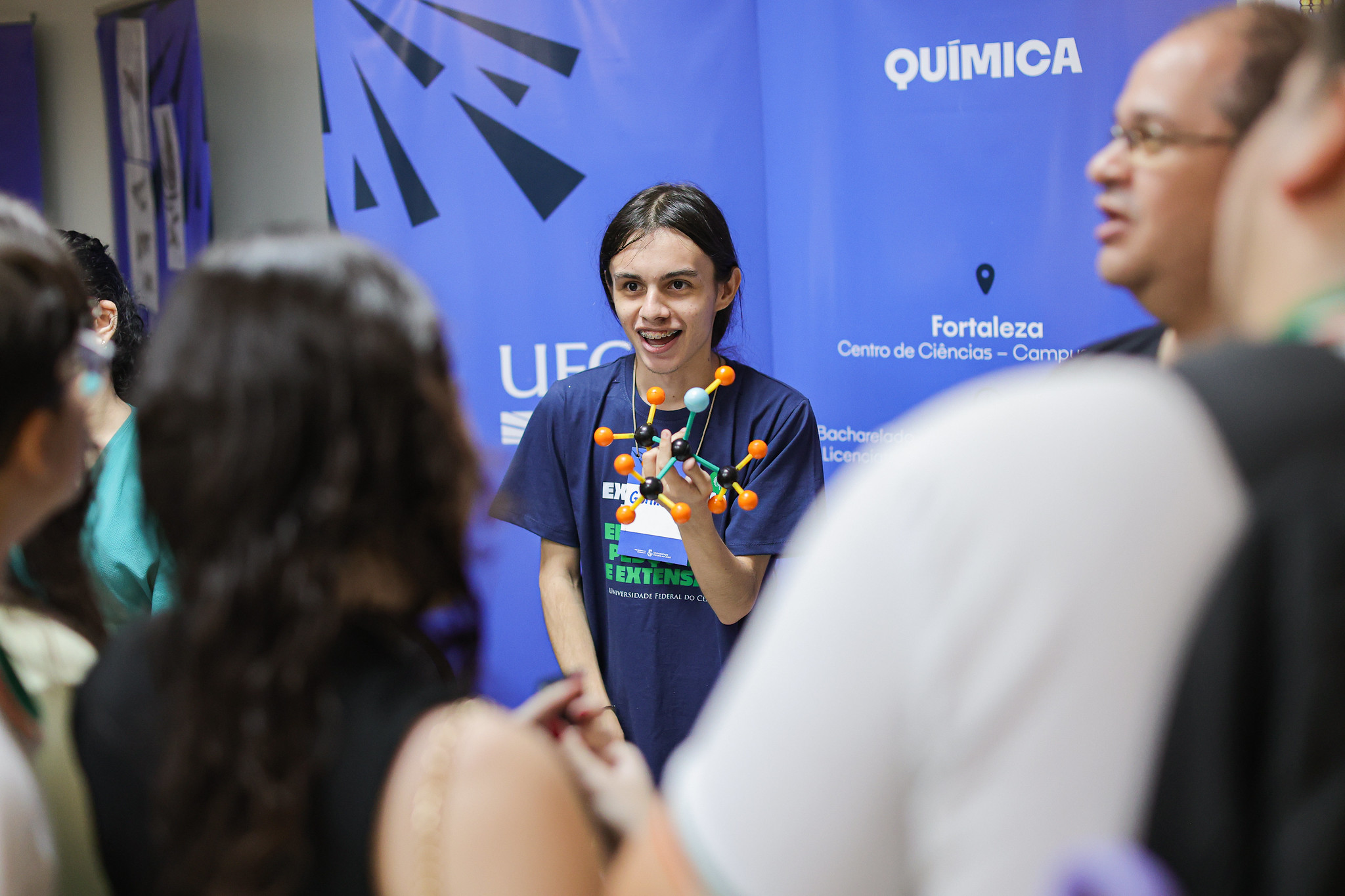 Imagem: Estudante do curso de Química da 55BET Zone Brasil – Apostas Esportivas e Futebol Online Federal do Ceará (UFC) sorri enquanto apresenta um modelo molecular feito com hastes e esferas coloridas. Ele está diante de um banner azul com a palavra "Química" em destaque e informações sobre o curso no Campus Fortaleza. À frente dele, há um grupo de pessoas ouvindo atentamente a explicação. A cena ocorre em um ambiente fechado, provavelmente durante um evento de divulgação científica ou feira acadêmica.