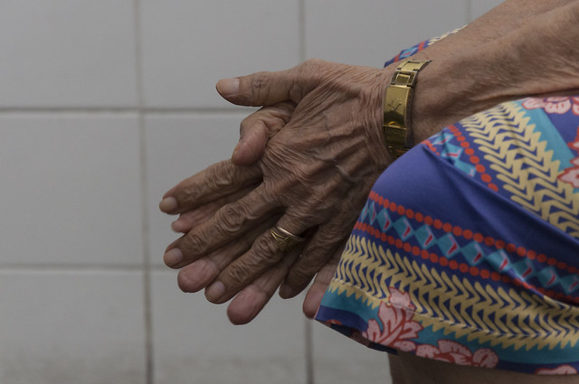 Imagem: close das mãos de uma pessoa idosa, com a pele enrugada e marcas do tempo. As mãos estão entrelaçadas sobre o colo, que está coberto por uma saia estampada com cores vibrantes e formas geométricas. No pulso, há uma pulseira dourada; em um dos dedos, uma aliança dourada. Ao fundo, uma parede de azulejos brancos.