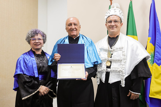 Imagem: três pessoas estão em pé, sorrindo, em um ambiente interno. Ao centro, um homem idoso, de pele clara e calvo, veste beca preta com faixa azul e segura um diploma emoldurado com as duas mãos. À esquerda, uma mulher de pele clara, cabelo cacheado grisalho e óculos, veste beca preta com faixa azul. À direita, um homem de pele clara, usa óculos e veste beca preta com capa branca e um chapéu cerimonial branco, com adornos dourados. Ao fundo, as bandeiras do Brasil e do estado do Ceará.