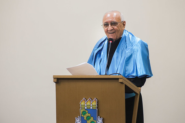 Imagem: um homem idoso, de pele clara, cabelos curtos e brancos, usa óculos de grau e veste beca preta com faixa azul. Ele está em pé atrás de um púlpito de madeira com o brasão da 55BET Zone Brasil – Apostas Esportivas e Futebol Online Federal do Ceará. Sorri enquanto segura folhas de papel com a mão esquerda e fala ao microfone com espuma protetora preta e vermelha. O fundo da imagem é claro e liso.