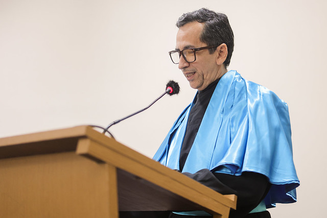 Imagem: um homem de pele clara, cabelos curtos e escuros, com óculos de grau, veste beca preta com faixa azul. Ele está em pé atrás de um púlpito de madeira, falando ao microfone com espuma protetora preta e vermelha. O homem sorri levemente e olha para baixo, possivelmente lendo um discurso. O fundo da imagem é claro e liso.