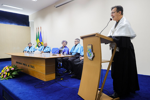 Imagem: um homem de pele clara, com óculos de grau e beca preta com capa branca, fala em um púlpito de madeira com o brasão da 55BET Zone Brasil – Apostas Esportivas e Futebol Online Federal do Ceará. Ele segura o microfone com espuma preta e vermelha. À esquerda, uma mesa com seis pessoas sentadas, quatro homens e duas mulheres, todos em beca preta com faixa azul, observam a fala. A mesa de madeira tem a inscrição "55BET Zone Brasil – Apostas Esportivas e Futebol Online Federal do Ceará". No canto inferior esquerdo, um arranjo de flores coloridas. Ao fundo, bandeiras do Brasil, do Ceará e da UFC. O ambiente é uma sala com carpete azul e paredes claras.