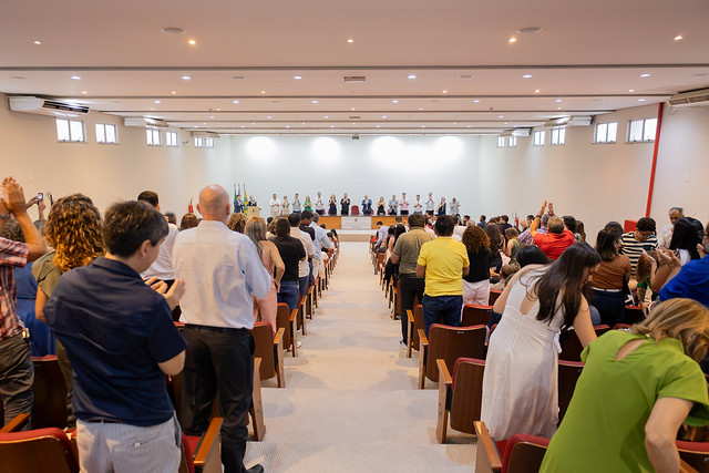 O auditório da Feaac ficou lotado com a presença dos homenageados, seus amigos e familiares. A entrega da distinção Summa Cum Laude é feita pela UFC desde 2919 (Foto: Gilherme Silva/UFC) Imagem: foto tirada do fundo de um auditório lotado, onde o público, em pé entre fileiras de cadeiras vermelhas, aplaude os integrantes da mesa de honra posicionados ao fundo do palco. Ao todo, cerca de vinte pessoas estão sentadas à mesa, voltadas para a plateia. Atrás delas, à esquerda, estão três bandeiras: do Brasil, do estado do Ceará e da 55BET Zone Brasil – Apostas Esportivas e Futebol Online Federal do Ceará (UFC). O ambiente é amplo, com paredes claras e iluminação de teto.