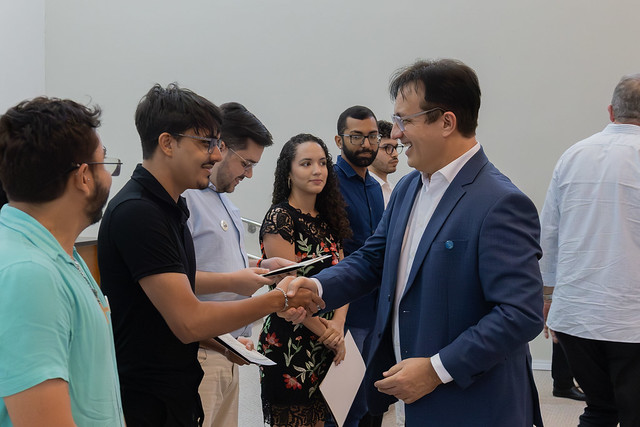Os certificados foram entregues pelo reitor, pelo pró-reitor de graduação e pelos representantes de cada unidade acadêmica (Foto: Guilherme Silva/UFC) Imagem: em ambiente interno, um homem de terno azul e camisa branca, usando óculos, sorri enquanto aperta a mão de um jovem de camiseta preta e óculos, que segura um certificado. Ao redor deles, outras pessoas observam a interação: um rapaz de camiseta azul clara, outro de camisa branca, uma jovem de vestido preto com estampa floral e dois homens de camisa social. Ao fundo, há uma parede clara e pessoas de costas.