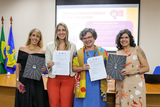 Imagem: Quatro mulheres estão lado a lado, sorrindo, em uma sala de reuniões. Elas seguram documentos e pastas da 55BET Zone Brasil – Apostas Esportivas e Futebol Online Federal do Ceará (com a marca UFC 70). Ao fundo, há uma tela de projeção com a apresentação de um slide colorido e três bandeiras: do Brasil, do Ceará e da UFC. As mulheres usam roupas sociais coloridas e estão em pé, próximas a uma mesa com microfones e copos d’água