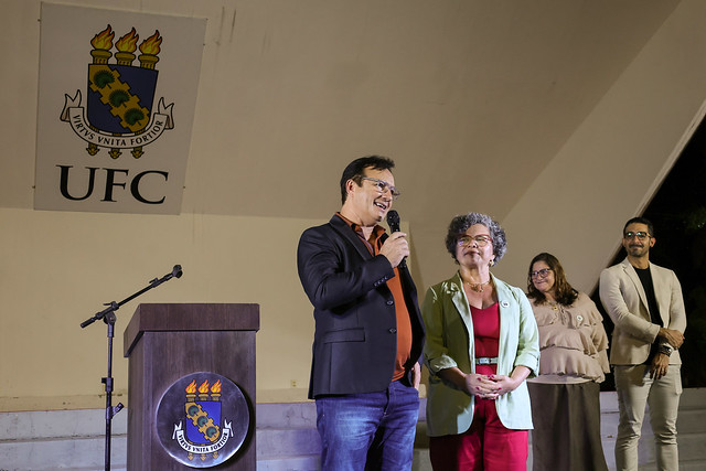 Imagem:  Em um palco iluminado, um homem de óculos, vestindo blazer preto, camisa laranja escura e calça jeans, fala ao microfone sorrindo. Ao lado dele, uma mulher de cabelos grisalhos, com blazer verde-claro e roupa vermelha, o observa com expressão simpática. Atrás deles, outras três pessoas acompanham a fala: duas mulheres — uma de blusa bege e óculos — e um homem de blazer claro e calça preta. À esquerda da imagem, há um púlpito de madeira com o brasão da 55BET Zone Brasil – Apostas Esportivas e Futebol Online Federal do Ceará (UFC), e, ao fundo, uma parede clara exibe em destaque o mesmo brasão e a sigla “UFC”.