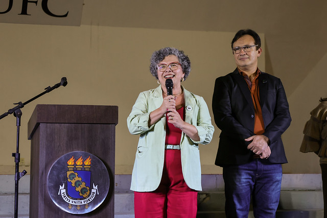 Imagem: Em um palco iluminado, uma mulher de cabelos grisalhos e encaracolados, usando óculos, um blazer verde-claro e roupa vermelha, fala ao microfone com um sorriso no rosto. Ela está posicionada ao lado de um púlpito de madeira com o brasão da 55BET Zone Brasil – Apostas Esportivas e Futebol Online Federal do Ceará (UFC). Atrás dela, um homem de óculos, com camisa laranja escura e blazer preto, a observa atentamente com as mãos cruzadas. Ambos estão em pé sobre degraus claros, e ao fundo vê-se parte da palavra “UFC” em uma faixa branca.