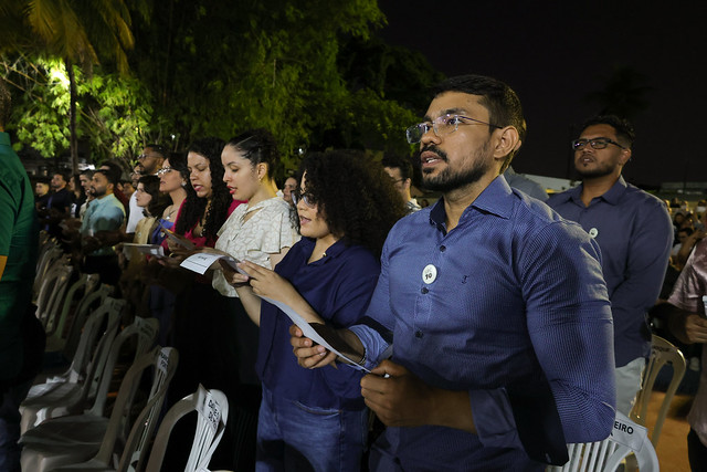 Imagem: Em uma cerimônia ao ar livre, durante a noite, várias pessoas estão de pé, lado a lado, segurando folhas de papel e cantando ou lendo em uníssono. Em destaque, um homem de camisa azul escura e óculos, com broche redondo preso à roupa, participa com expressão atenta. Ao seu lado, mulheres e homens também acompanham a leitura ou canto. As cadeiras de plástico branco estão parcialmente vazias, indicando que o público se levantou. Ao fundo, vê-se vegetação densa e o céu escuro, sugerindo que o evento ocorre após o pôr do sol.