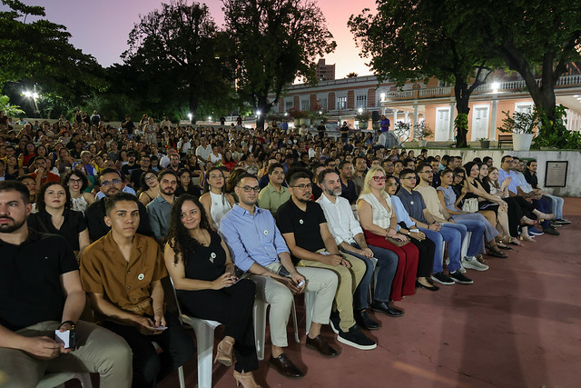 Imagem: Em um espaço ao ar livre, durante o entardecer, centenas de pessoas estão sentadas em uma concha acústica, voltadas para um palco fora do campo de visão. Na primeira fila, há homens e mulheres adultos, diversos em aparência e vestimentas, com expressões serenas e atentas. Muitos usam um broche circular claro preso à roupa. Atrás deles, os degraus da arquibancada estão completamente ocupados por um público numeroso. Ao fundo, vê-se um prédio histórico iluminado, com paredes em tom salmão e janelas brancas, cercado por árvores altas.