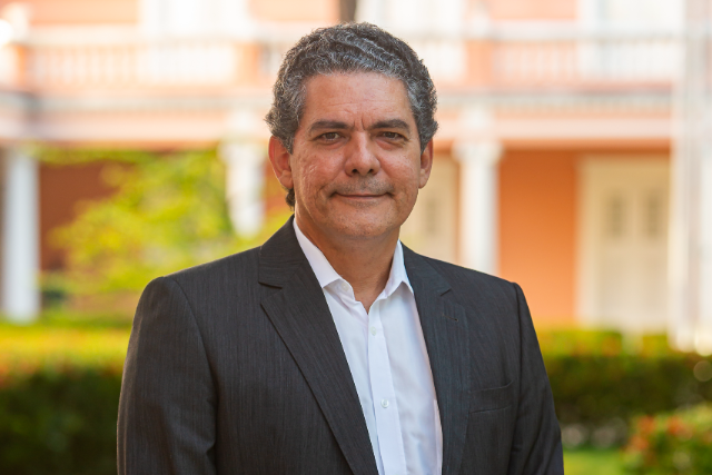 Joaquim Perúcio é o novo diretor-presidente da Fastef, fundação de apoio institucional à UFC (Foto: Nicolás Leiva) Imagem: Em ambiente externo e iluminado pela luz natural, um homem de pele clara, cabelos grisalhos ondulados e expressão serena está posando para a foto. Ele veste paletó escuro e camisa social branca, sem gravata. Ao fundo, desfocado, vê-se um prédio de fachada em tom salmão com colunas e sacadas brancas, além de vegetação bem cuidada, indicando um ambiente institucional ou histórico.