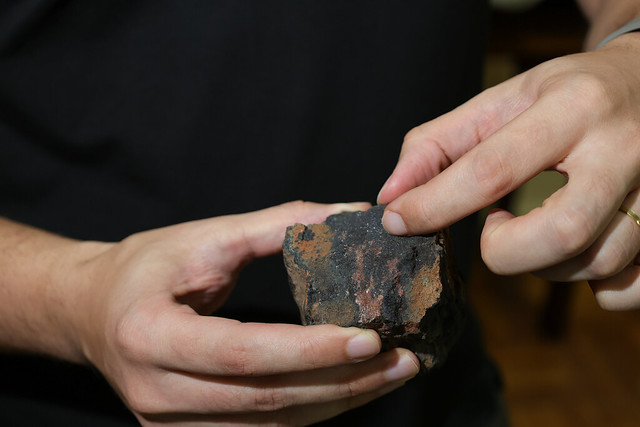 Um dos objetivos do seminário é evidenciar a importância da UFC para o desenvolvimento sustentável da mineração no Ceará, por meio de cursos como Engenharia de Minas e Geologia (Foto: Viktor Braga/UFC) Imagem: duas mãos seguram um pedaço irregular de rocha escura, com manchas avermelhadas e aspecto poroso. Um dos dedos da pessoa aponta para a superfície da pedra, destacando sua textura e detalhes minerais.