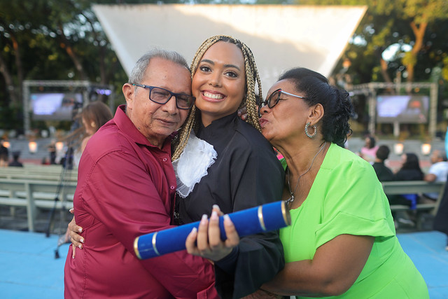 Imagem: jovem negra sorridente veste beca preta com faixa azul e segura o canudo de formatura ao lado de um homem e uma mulher mais velhos, que parecem ser seus familiares. O homem usa camisa vermelha e a mulher está com vestido verde-limão. Eles posam juntos em frente a um palco branco ao ar livre, com outras pessoas ao fundo participando da cerimônia de colação de grau.