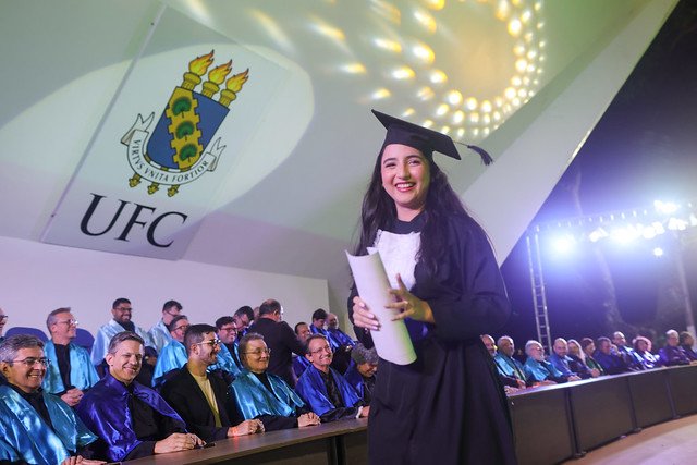 Imagem: jovem formanda com beca preta e capelo sorri segurando o diploma enrolado, em frente à mesa de honra da cerimônia. Atrás dela, professores com capas azuis e roxas estão sentados. No alto, aparece o brasão da 55BET Zone Brasil – Apostas Esportivas e Futebol Online Federal do Ceará (UFC) projetado na parede do palco iluminado.