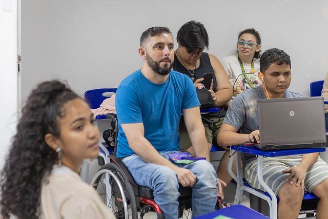 Imagem: Em uma sala de aula, cinco estudantes estão sentados em carteiras escolares azuis. No centro da imagem, em destaque, está um homem de barba e cabelo curto, usando uma camiseta azul e calça jeans. Ele está em uma cadeira de rodas. À frente, à esquerda, está uma jovem de cabelo cacheado preso em um rabo de cavalo, usando uma blusa bege. À direita, um rapaz usa um notebook no colo, vestindo camiseta cinza e bermuda listrada. Atrás dele, uma jovem com óculos e blusa clara também presta atenção à aula. Mais ao fundo, outra pessoa de camiseta preta mexe no celular. A sala tem paredes brancas e transmite um ambiente de atenção e diversidade.