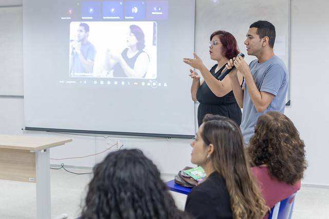 Imagem: Em uma sala de aula, dois intérpretes de Libras (Língua Brasileira de Sinais) estão em pé, lado a lado, em frente a uma tela de projeção. A mulher, de cabelo curto e vermelho, usa óculos e vestido preto; ela está sinalizando com as mãos. Ao seu lado, um homem jovem, com cabelo curto e camiseta azul, também sinaliza. Na tela ao fundo, é projetada uma videoconferência, mostrando a imagem de dois outros intérpretes em preto e branco. À frente, sentados, há quatro estudantes de costas, assistindo à apresentação.
