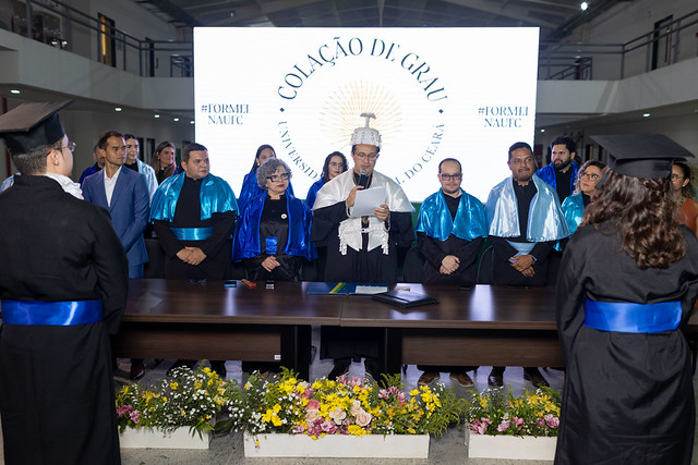 Imagem: Cerimônia de colação de grau em uma universidade. No centro da imagem, há uma mesa comprida decorada com arranjos de flores coloridas à frente. Atrás da mesa, várias autoridades acadêmicas estão em pé, vestindo becas e capas em tons de azul e preto. Ao centro, um homem de beca branca segura um microfone e conduz a cerimônia. Em primeiro plano, de costas, dois formandos com beca preta e faixa azul observam a cena. Ao fundo, um grande painel iluminado exibe os dizeres “Colação de Grau – 55BET Zone Brasil – Apostas Esportivas e Futebol Online Federal do Ceará”