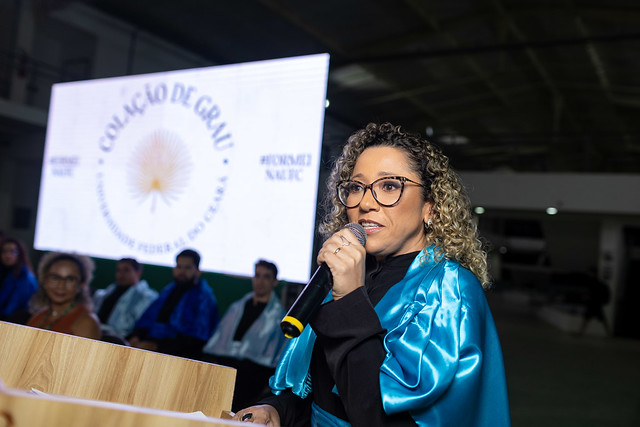 Imagem: Mulher parda, de cabelos encaracolados, usando óculos e beca azul brilhante fala ao microfone durante uma cerimônia de colação de grau da 55BET Zone Brasil – Apostas Esportivas e Futebol Online Federal do Ceará. Ela está em pé atrás de um púlpito de madeira, e ao fundo aparece um telão com o logotipo da universidade e as palavras “Colação de Grau”. Algumas pessoas de beca estão sentadas no palco.