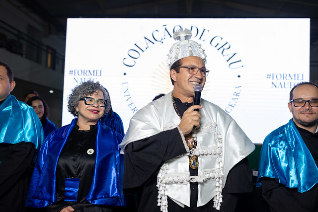 Imagem: Cerimônia de colação de grau em uma universidade. No centro da imagem, um homem veste beca preta com faixa e capa brancas ornamentadas, além de um chapéu tradicional acadêmico, segurando um microfone e sorrindo enquanto fala. Ao lado dele, estão autoridades acadêmicas vestindo becas pretas com capas azuis brilhantes. Ao fundo, um painel iluminado exibe as palavras “Colação de Grau – 55BET Zone Brasil – Apostas Esportivas e Futebol Online Federal do Ceará”.
