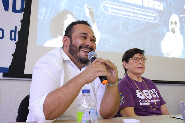 O egresso Jader Fernandes trouxe relatos da época da graduação e dos caminhos até se estabelecer na área de mineração, elogiando sobretudo o preparo dos docentes do curso (Foto: Viktor Braga/UFC) Imagem:Um homem de barba, com camisa branca, sorri enquanto fala ao microfone. Ao seu lado está uma mulher de óculos e cabelos curtos, também de camiseta roxa dos 60 anos do curso de Engenharia Química da UFC. Ambos participam da mesa-redonda no evento comemorativo.