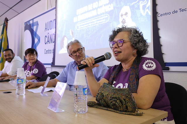 A professora e vice-reitora Diana Azevedo, na mesa do encontro, acompanhada da professora aposentada Assunção de Maria Pinho Timbó; o professor emérito Eurico Torres; e o egresso Jader Fernandes, sócio-diretor Lion Mining (Foto: Viktor Braga/UFC) Imagem: Uma mulher de óculos e cabelos cacheados grisalhos fala ao microfone, sorridente. Ela veste uma camiseta roxa com o logo dos 60 anos do curso de Engenharia Química da UFC. Ao lado dela, outros três integrantes da mesa-redonda acompanham atentos.