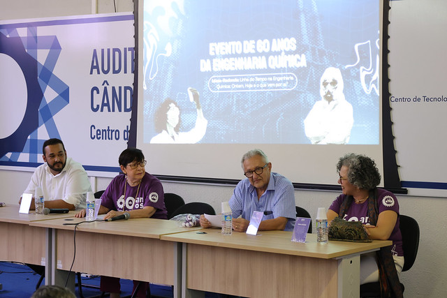 Reunindo professores, alunos e egressos, o encontro rendeu conversas sobre a história, o legado e os caminhos para o futuro do curso de Engenharia Química da UFC, além de perspectivas para a profissão (Foto: Viktor Braga/UFC) Imagem: Quatro pessoas estão sentadas atrás de uma mesa em um auditório, participando de uma mesa-redonda. Duas mulheres usam camisetas roxas com o logo dos 60 anos do curso de Engenharia Química da UFC, enquanto um homem de camisa azul clara e outro de camisa branca também compõem a mesa. Ao fundo, um telão exibe a mensagem “Evento de 60 anos da Engenharia Química”.