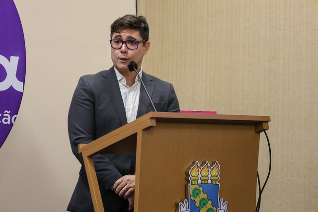 Imagem: Homem jovem, de terno cinza e óculos, fala ao microfone em púlpito com o brasão da UFC. Ao fundo, parte do painel roxo com o logotipo da UFC Inova aparece.