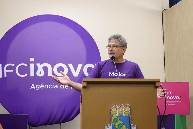 Imagem: Homem de cabelos grisalhos e óculos fala em púlpito de madeira com o brasão da UFC. Ele veste camiseta roxa com a palavra “Maior”. Atrás dele há um grande painel circular roxo com o logotipo da Agência UFC Inova.