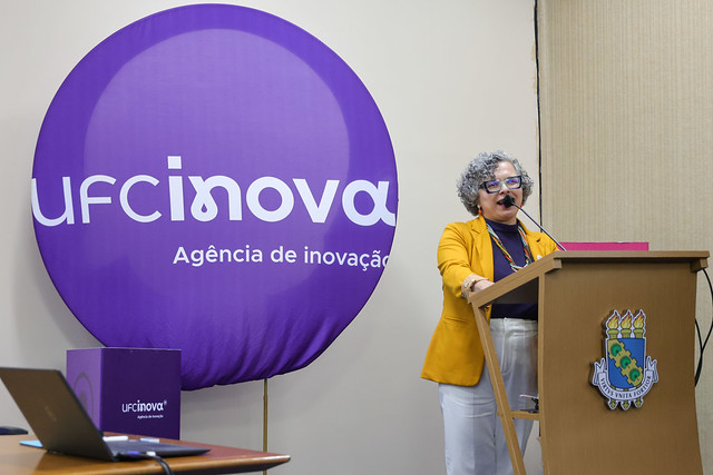 Imagem: Mulher de cabelos cacheados grisalhos, óculos e batom vermelho fala ao microfone em púlpito com o brasão da UFC. Ela veste casaco mostarda e blusa roxa. Ao fundo, um grande painel roxo exibe o logotipo da Agência UFC Inova.