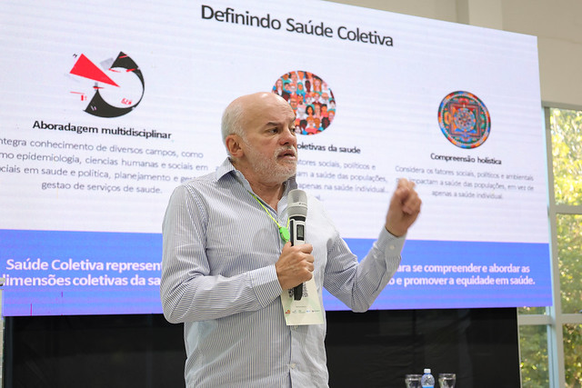 Na conferência de abertura, o pesquisador da Fiocruz Rômulo Paes de Sousa abordou a relação entre saúde coletiva, meio ambiente e sustentabilidade (Foto: Viktor Braga/UFC) Imagem: Homem de cabelos e barba brancos fala ao microfone durante apresentação em auditório. Ele usa camisa listrada azul clara e crachá no pescoço. Atrás dele, um telão exibe o slide com o título “Definindo Saúde Coletiva” e três tópicos com ícones ilustrativos sobre abordagem multidisciplinar, dimensões coletivas da saúde e compreensão holística. O ambiente é iluminado e há janelas amplas ao fundo, com vista para árvores.