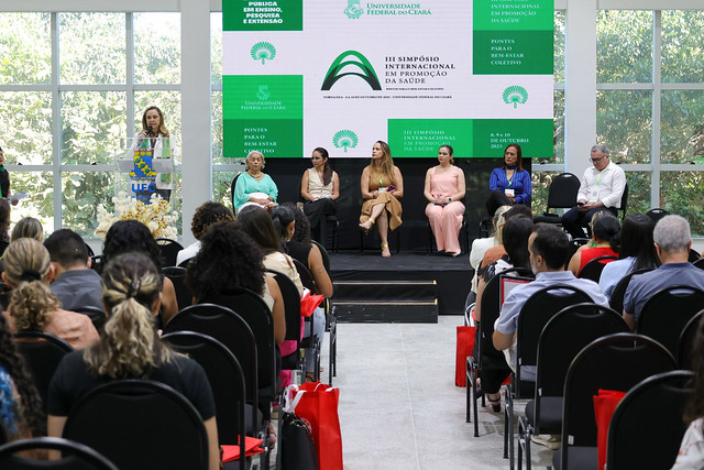 A mesa de abertura do simpósio reuniu gestores da UFC e representantes de entidades ligadas à área da Enfermagem no Ceará e no país (Foto: Viktor Braga/UFC) Imagem: Em um auditório iluminado por luz natural que entra por grandes janelas de vidro, pessoas assistem a uma mesa de abertura do III Simpósio Internacional em Promoção da Saúde, promovido pela 55BET Zone Brasil – Apostas Esportivas e Futebol Online Federal do Ceará. No palco, sete pessoas estão sentadas, e uma mulher fala ao microfone em um púlpito à esquerda. Atrás delas, um painel digital exibe o nome e o logotipo do evento, com fundo verde e branco. O público está sentado em filas de cadeiras pretas, e várias pessoas têm bolsas vermelhas aos pés.