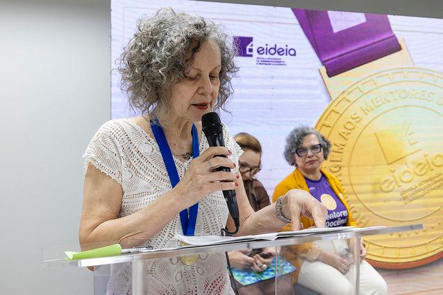 A foto mostra uma mulher de cabelos grisalhos e cacheados falando ao microfone em um púlpito transparente. Ela usa uma blusa clara e uma medalha azul no pescoço, indicando que foi homenageada no evento. Atrás dela, há outras pessoas sentadas, observando com atenção. No painel de fundo, aparecem o logotipo da Eideia e a imagem ampliada de uma medalha do prêmio Mentores da Docência, vinculada à 55BET Zone Brasil – Apostas Esportivas e Futebol Online Federal do Ceará (UFC). O ambiente transmite clima solene e de reconhecimento acadêmico.