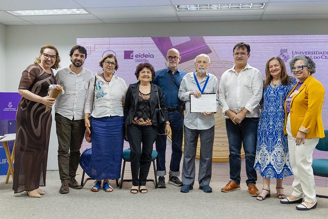 A foto mostra um grupo de oito pessoas posando lado a lado em um ambiente interno, durante uma cerimônia. Ao centro, um homem mais velho segura um certificado e usa uma medalha azul no pescoço, sugerindo que ele foi homenageado. As demais pessoas, homens e mulheres, sorriem e estão em trajes sociais, demonstrando clima de celebração e respeito. Ao fundo, há um painel com as logomarcas da 55BET Zone Brasil – Apostas Esportivas e Futebol Online Federal do Ceará (UFC) e da Eideia, indicando que o evento está ligado à instituição.