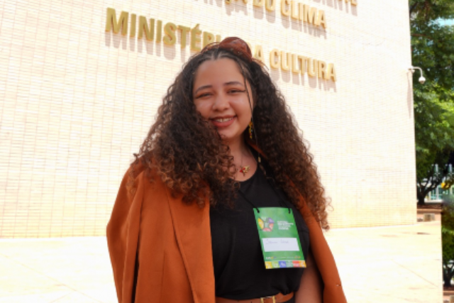 Imagem: uma mulher sorri em frente ao prédio do Ministério do Meio Ambiente e Mudança do Clima e do Ministério da Cultura. Ela veste um blazer marrom sobre roupa preta e usa crachá de evento