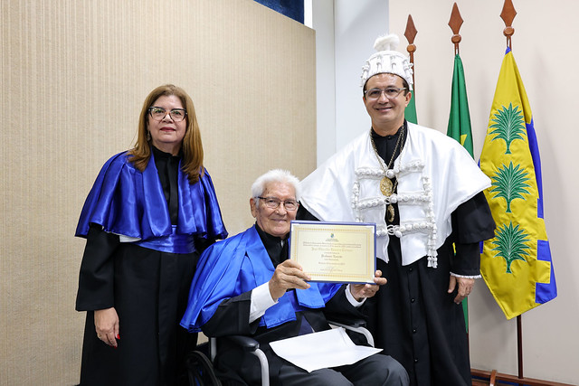 Imagem: Uma mulher com beca azul e um homem com traje cerimonial branco posam ao lado de uma pessoa idosa sentada em cadeira de rodas, que segura um certificado. As bandeiras oficiais estão posicionadas atrás deles.