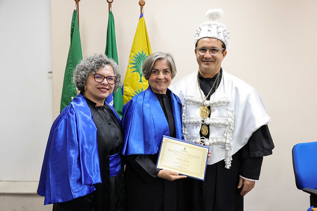 Imagem: Duas mulheres com becas pretas e estolas azuis posam ao lado de um homem com traje branco cerimonial detalhado e um chapéu tradicional. A pessoa ao centro segura um certificado. Bandeiras oficiais aparecem ao fundo.