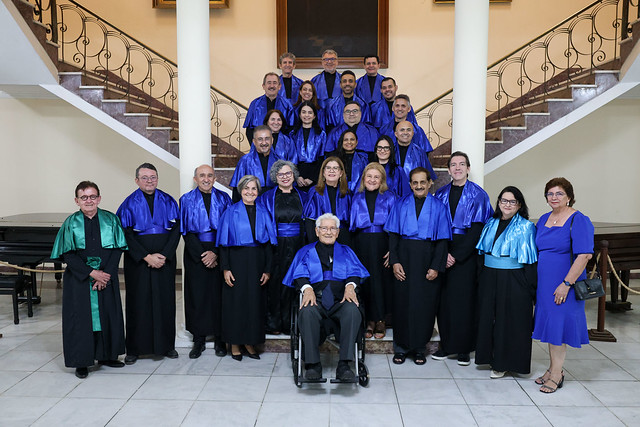 Imagem: Grupo grande de pessoas posando para foto em um hall com escadaria ao fundo. Todos usam becas pretas com estolas azuis, exceto uma pessoa com estola verde. No centro da primeira fileira, uma pessoa idosa usa a beca azul enquanto está sentada em uma cadeira de rodas.