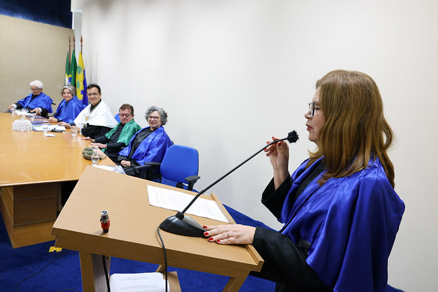 Imagem: Mulher usando beca azul fala ao microfone em um púlpito de madeira, segurando um papel. À sua frente, uma mesa comprida onde várias pessoas, também com becas coloridas, estão sentadas observando. Ao fundo, bandeiras oficiais estão posicionadas no canto da sala.