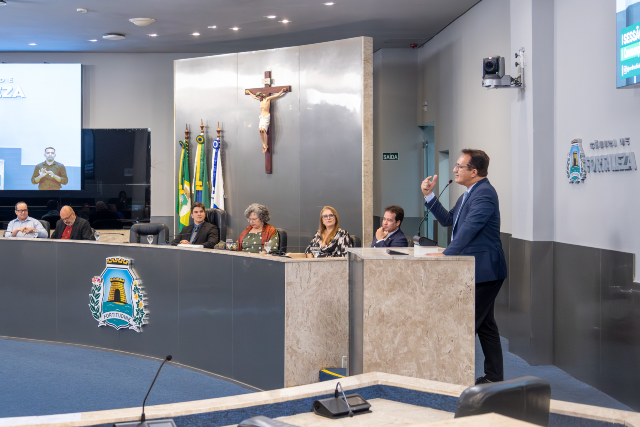 Imagem: vista lateral do plenário da Câmara de Fortaleza. Um homem de terno escuro está em pé, discursando e gesticulando com uma das mãos. À sua frente, uma mesa elevada em formato semicircular reúne outras pessoas sentadas, ouvindo atentamente. Ao fundo, há um crucifixo na parede, bandeiras oficiais e telas de transmissão. O ambiente é amplo, com iluminação clara e institucional.