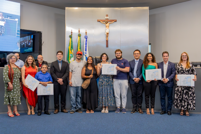 Imagem: grupo de cerca de 14 pessoas, entre homens e mulheres de diferentes idades, tons de pele e estilos de roupa, posam em pé em um plenário. Algumas seguram certificados. Ao fundo, há um grande crucifixo fixado na parede, bandeiras do Brasil, do Ceará e de Fortaleza, e o espaço típico de uma câmara legislativa. Todos estão alinhados lado a lado, voltados para a câmera.