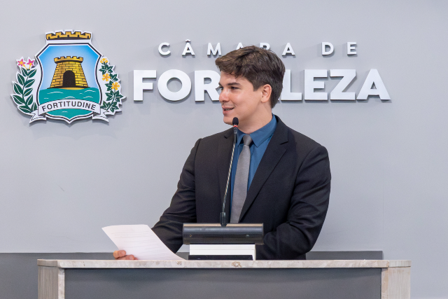 Imagem: homem jovem, de pele clara e cabelo castanho curto, veste terno escuro, camisa azul e gravata. Ele está em pé atrás de um púlpito, falando ao microfone e segurando um papel. Ao fundo, na parede clara, aparece o brasão e o nome “Câmara de Fortaleza”. O homem olha levemente para o lado, com expressão atenta e concentrada.