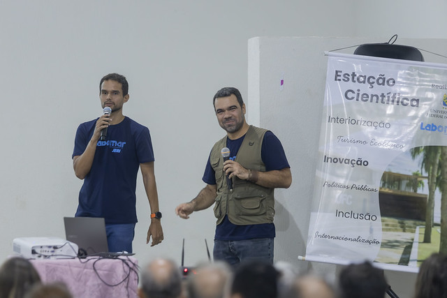 O professor do Labomar Marcelo Soares (direita) e o superintendente de infraestrutura da UFC, Renato Guerreiro, durante apresentação do projeto (Foto: Guilherme Silva/UFC) Imagem: Dois homens estão em pé em um auditório, falando ao público com microfones. À esquerda, um homem jovem veste camiseta azul escura e segura um microfone com a mão direita, ao lado de uma mesa com notebook e projetor. À direita, outro homem, também jovem, usa camiseta azul e um colete verde com bolsos, segurando um microfone e sorrindo levemente. Ao fundo, há um banner vertical com o título “Estação Científica” e palavras como “Interiorização”, “Inovação”, “Políticas Públicas”, “Inclusão” e “Internacionalização”. Em primeiro plano, aparecem as cabeças do público sentado, visto de costas.