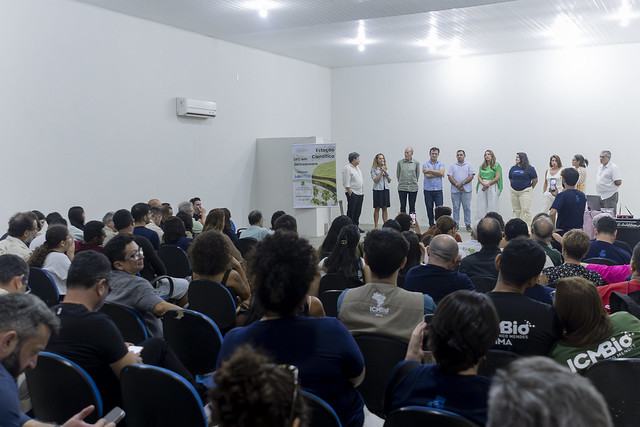 Auditório lotado na cerimônia que oficializou o início das obras da Estação Científica: evento marca chegada física da UFC no litoral oeste do Ceará, em um projeto sonhado coletivamente (Foto: Guilherme Silva/UFC) Imagem: Auditório cheio, com dezenas de pessoas sentadas em cadeiras pretas, voltadas para a frente. No palco, um grupo de homens e mulheres está alinhado lado a lado. Uma mulher fala ao microfone enquanto os demais escutam. O espaço é amplo, com paredes brancas e iluminação no teto.