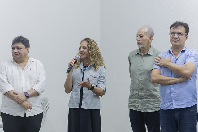 A gestora do Parque Nacional de Jericoacoara, Kelly Cottens, comemorou, entre outros aspectos, a previsão do funcionamento de um curso de graduação em Turismo Ecológico na Estação Científica (Foto: Guilherme Silva/UFC) Imagem: quatro pessoas estão em pé diante de uma parede branca. Ao centro, uma mulher de cabelo cacheado e claro segura um microfone e fala, com expressão confiante. À esquerda e à direita dela, três homens observam atentamente. Todos vestem roupas casuais, em um ambiente interno bem iluminado.