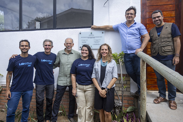 Placa do posto de cooperação técnico-operacional da UFC na sede do ICMBio: estrutura para receber pesquisadores e estudantes e para desenvolver ações relacionadas ao Parque Nacional de Jericoacoara (Foto: Guilherme Silva/UFC) Imagem: grupo de oito pessoas posa para foto em frente a um prédio institucional. Sete pessoas estão alinhadas em pé, lado a lado, sorrindo para a câmera, e uma pessoa está apoiada em um corrimão à direita, em posição mais elevada. Algumas pessoas vestem camisetas azul-escuro com a palavra “Labomar” escrita em azul claro. No centro do grupo, há um homem mais velho de camisa verde clara. Ao fundo, na parede do prédio, vê-se uma placa comemorativa fixada. O ambiente externo inclui parede branca, detalhes em tijolo, plantas e um pequeno jardim na parte inferior da imagem.
