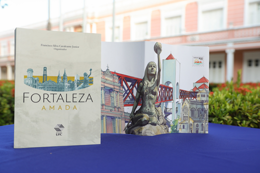 O livro Fortaleza Amada já pode ser adquirido na Editora da UFC; os 100 primeiros compradores levarão como brinde um planner na mesma identidade visual da obra. (Foto: Viktor Braga/ UFC Informa) Imagem: Foto do livro Fortaleza Amada sobre uma mesa; ao fundo, o prédio da Reitoria