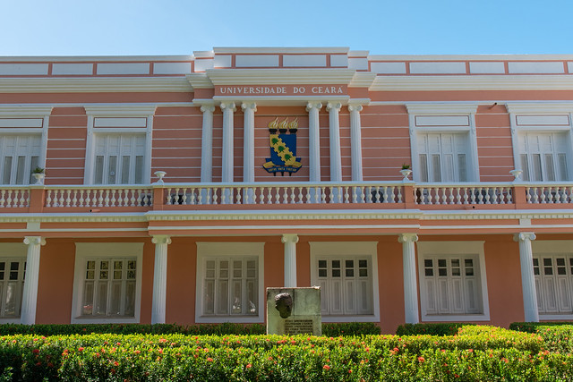 Imagem: fachada do prédio da Reitoria da UFC, com paredes na cor rosa e colunas e janelas brancas. Na frente da fachada, há um gramado e um monumento com a escultura de rosto em bronze