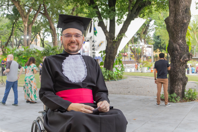 Imagem: Homem jovem com barba curta e óculos usa beca preta, faixa vermelha na cintura e capelo. Ele está sentado em uma cadeira de rodas, sorrindo para a câmera, em espaço arborizado ao ar livre. Ao fundo, há árvores grandes, palco montado e pessoas caminhando, reforçando o clima de celebração da cerimônia de formatura.