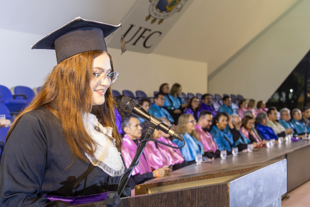 Imagem: Jovem mulher com cabelos longos ruivos usa beca preta, faixa roxa na cintura e capelo (chapéu de formatura). Ela sorri levemente enquanto lê um texto ao microfone em um púlpito de madeira. Ao fundo, autoridades acadêmicas vestidas com becas coloridas acompanham a cerimônia, sentadas em uma longa mesa