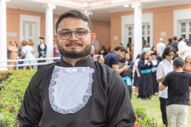 Imagem: Homem jovem, de barba e bigode aparados, usa óculos de armação escura e veste beca preta com gola branca rendada no peito. Ele está em pé, olhando diretamente para a câmera com expressão serena e leve sorriso. Ao fundo, em área externa com jardim e prédio de fachada em tom salmão e branco, outras pessoas circulam e conversam, indicando ambiente de formatura